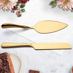 May include: A set of two gold-colored cake servers. The larger server has a serrated edge and a slightly curved blade. The smaller server has a straight blade. Both servers have long, slender handles.