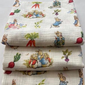 Peut inclure: Une pile de quatre couvertures en mousseline pliées présentant un motif répétitif de personnages de Pierre Lapin, de carottes orange, de radis rouges et de feuilles vertes sur un fond blanc cassé. Le tissu a une texture douce et froissée.