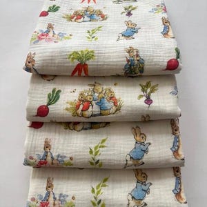 Puede incluir: Cuatro mantas de muselina blanca dobladas con un patrón repetido de personajes de Peter Rabbit, zanahorias y rábanos. Las mantas están apiladas, mostrando las ilustraciones detalladas en colores suaves.