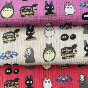 Può includere: Tre strati di tessuto in rosa, crema e rosso, ciascuno con motivi di personaggi dei cartoni animati. I disegni includono una grande creatura grigia e rotonda, un gatto nero, un autobus con la faccia di un gatto e altre figure stravaganti. Il tessuto sembra morbido e strutturato.