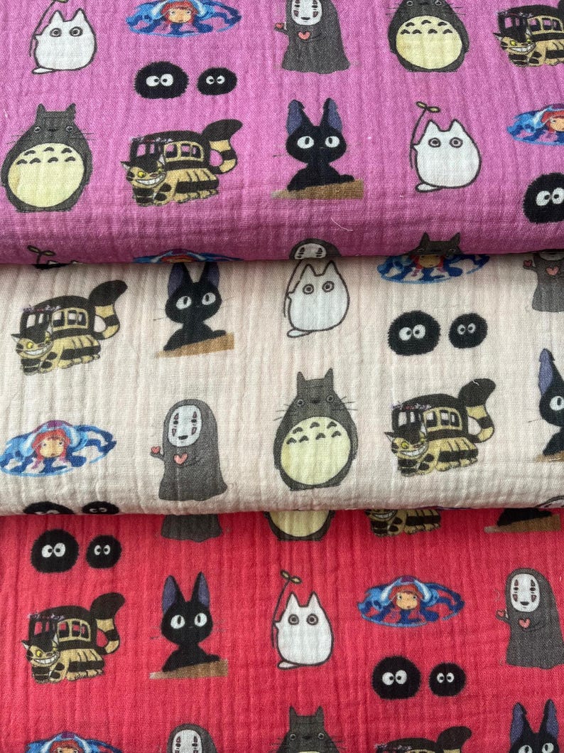 Puede incluir: Tres capas de tela en rosa, crema y rojo, cada una estampada con personajes de dibujos animados. Los dise&ntilde;os incluyen una criatura grande, redonda y gris, un gato negro, un autob&uacute;s con cara de gato y otras figuras caprichosas. La tela parece suave y texturizada.