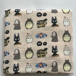 Puede incluir: Tela beige doblada con un patr&oacute;n repetido de personajes animados. El dise&ntilde;o incluye im&aacute;genes de una criatura grande, gris y peluda, un gato negro, un gato blanco, un autob&uacute;s y una figura enmascarada.