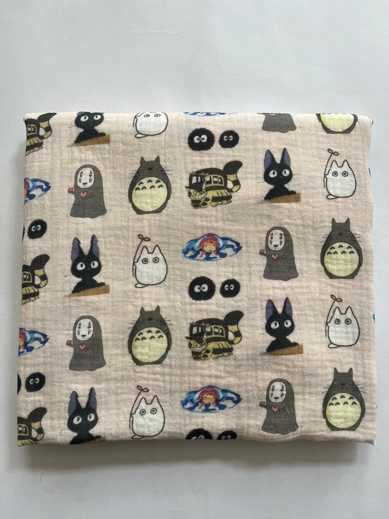 Puede incluir: Tela beige doblada con un patr&oacute;n repetido de personajes animados. El dise&ntilde;o incluye im&aacute;genes de una criatura grande, gris y peluda, un gato negro, un gato blanco, un autob&uacute;s y una figura enmascarada.