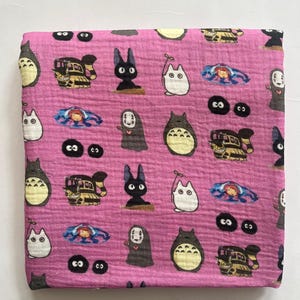 Puede incluir: Tela rosa con un patr&oacute;n repetido de personajes animados. El dise&ntilde;o incluye im&aacute;genes de una criatura grande, redonda y gris con vientre blanco, un gato negro y un tren. Tambi&eacute;n hay otros personajes.