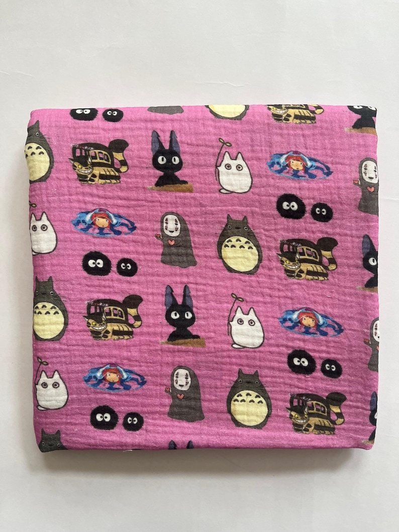 Puede incluir: Tela rosa con un patr&oacute;n repetido de personajes animados. El dise&ntilde;o incluye im&aacute;genes de una criatura grande, redonda y gris con vientre blanco, un gato negro y un tren. Tambi&eacute;n hay otros personajes.
