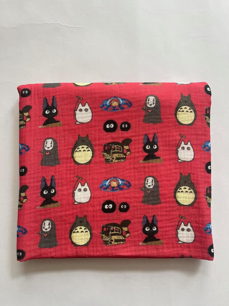 Puede incluir: Tela roja plegada con un estampado repetido de personajes de dibujos animados. El dise&ntilde;o incluye varias criaturas en negro, blanco y gris, con algunos toques de azul. La tela est&aacute; doblada, lo que sugiere que podr&iacute;a ser una bufanda o un accesorio similar.
