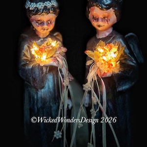 May include: Two dark angel figurines holding glowing floral bouquets. The angels have dark hair, crowns, and a distressed appearance. The bouquets emit a warm glow, and ribbons with small, clear beads hang down. The text "©Wicked Wonders Design 2026" is visible.