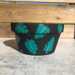 May include: A black terracotta planter with a teal leaf pattern. The planter is round and has a wide rim.