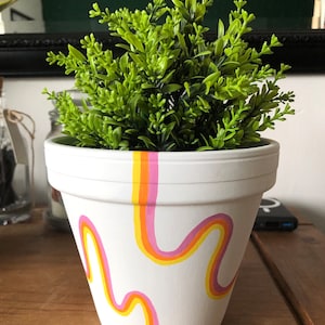 May include: White ceramic planter with a hand-painted design of wavy lines in pink, yellow, and orange. A green potted plant is inside the planter.