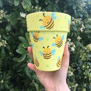 May include: A yellow ceramic planter with a bee pattern. The planter has a light yellow background with a repeating pattern of bees with black stripes and blue wings. The bees have smiling faces and pink cheeks.