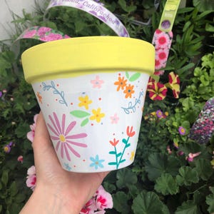 May include: A white ceramic plant pot with a yellow rim, adorned with vibrant floral patterns. The pot showcases pink, yellow, orange, and blue flowers, and green leaves. Ideal for planting flowers and other plants.