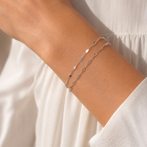 Op de afbeelding: Twee delicate zilverkleurige armbanden die samen worden gedragen. De ene armband heeft kleine, platte, reflecterende ovale schakels, terwijl de tweede armband bestaat uit een fijn, gedraaid kettingontwerp. Beide stukken hebben een glanzende afwerking.