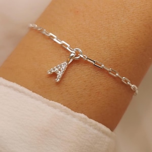 Op de afbeelding: Een zilverkleurige kettingarmband met een enkele letter A-bedel. De bedel is ingelegd met kleine, heldere, sprankelende steentjes. De armband bestaat uit langwerpige rechthoekige schakels die in een reeks met elkaar zijn verbonden.