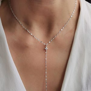 以下が含まれることがあります： Y字型のペンダントが付いた繊細なシルバーネックレス。ネックレスは、ペンダントの中央に小さな輝く宝石と、一連の小さな長方形のリンクが特徴です。白いシャツの上から着用されています。