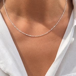 May include: A delicate silver chain necklace featuring small, reflective, flat circular links spaced evenly along the length of the chain. The necklace rests against the neck, showcasing a bright, metallic finish that catches the light.