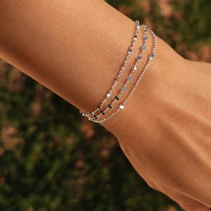 Op de afbeelding: Een drievoudige zilveren armband met fijne kettinkjes voorzien van kleine, platte, ronde metalen schijfjes die gelijkmatig over elke streng zijn verdeeld. De armband heeft een gepolijste, reflecterende afwerking en een gelaagd ontwerp.