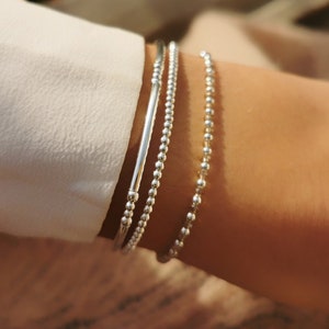 Può includere: Tre braccialetti d'argento sono impilati su un polso. Uno è una barra piena, gli altri due sono con perline. I braccialetti sono indossati sopra una camicia bianca a maniche lunghe. I braccialetti sono semplici ed eleganti.