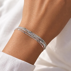 Könnte beinhalten: Ein mehrsträngiges Armband aus silberfarbenen Ketten mit kleinen, gleichmäßig verteilten runden Akzenten in verschiedenen Farben wie Blau, Rosa und Schwarz. Die feinen Metallketten sind übereinandergelegt und bilden ein schmales Armband.
