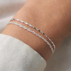 Könnte beinhalten: Ein zweireihiges Silberarmband bestehend aus einer Kette mit kleinen rechteckigen Gliedern und einer zweiten Kette mit gedrehtem Seilmuster. Die beiden feinen Metallketten werden zusammen getragen und haben eine helle, reflektierende Oberfläche.