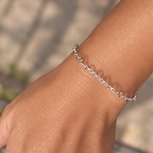 Op de afbeelding: Een zilveren kettingarmband bestaande uit een reeks kleine, ronde, getextureerde schakels. Het metaal heeft een heldere, reflecterende afwerking met een herhalend patroon van fijne, geëtste lijnen op elke individuele schakel.
