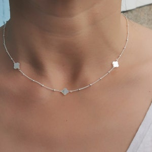 May include: A delicate silver necklace with four-leaf clover pendants. The necklace has a fine chain with small, round beads. The clover pendants are a light, pearly white colour. The necklace is worn around the neck.