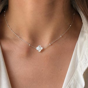 Op de afbeelding: Een zilverkleurige ketting met een centrale hanger in de vorm van een klavertjevier van iriserend wit parelmoer. De fijne ketting is voorzien van kleine, gelijkmatig verdeelde zilverkleurige kraaltjes die zorgen voor een subtiele textuur.