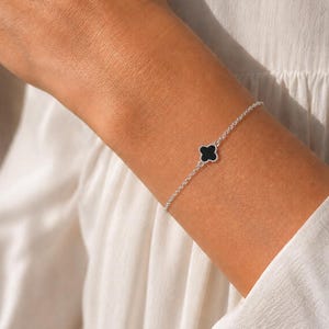 Puede incluir: Una delicada pulsera de cadena de plata con un dije central en forma de trébol de cuatro hojas. El dije de trébol tiene una incrustación negra lisa y sólida rodeada por un borde plateado fino. La pulsera presenta un diseño de cadena de metal.