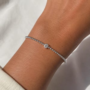 Peut inclure: Un bracelet délicat composé de petites perles argentées uniformes et polies, agrémenté d'une unique pierre claire ronde sertie dans un chaton circulaire en argent. Les perles sont enfilées pour former une bande continue et brillante.