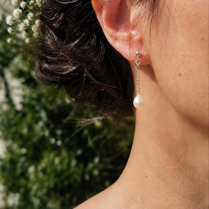 May include: A drop earring featuring a small, white, teardrop-shaped pearl suspended from a delicate silver-toned chain. The earring is attached to a simple silver-toned stud post that secures the piece to the earlobe, creating a subtle, dangling effect.