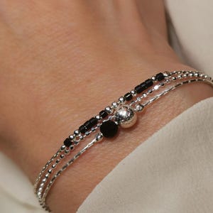 Könnte beinhalten: Drei zarte Silberarmbänder, die an einem Handgelenk getragen werden. Eines hat kleine schwarze Perlen, ein anderes eine einzelne schwarze Perle und eine silberne Kugel, und das dritte ist eine einfache Kette. Die Armbänder werden vor einem weißen Stoffhintergrund getragen.