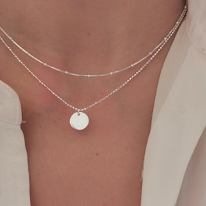 Puede incluir: Un collar de plata con una cadena delicada y un pequeño colgante redondo. El collar está superpuesto con una segunda cadena más fina.