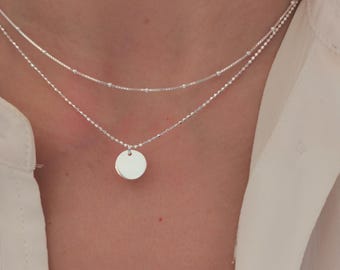 Op de afbeelding: Een zilveren ketting met een delicate ketting en een klein rond hangertje. De ketting is gelaagd met een tweede, dunnere ketting.
