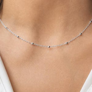 May include: A delicate silver-toned chain necklace featuring small, evenly spaced spherical beads along the length of the chain. The necklace rests against the neck, showcasing a simple, thin design with a polished metallic finish throughout.