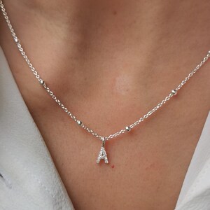 Peut inclure: Un collier argenté avec un petit pendentif en forme de lettre "A". Le pendentif est incrusté de petites pierres précieuses transparentes. La chaîne du collier comporte de petites perles rondes.