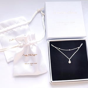 May include: A silver necklace with a small round pendant, packaged in a white box with a black interior and a white satin bag. The box and bag have the text "Solstice Blue Boutique" printed on them.