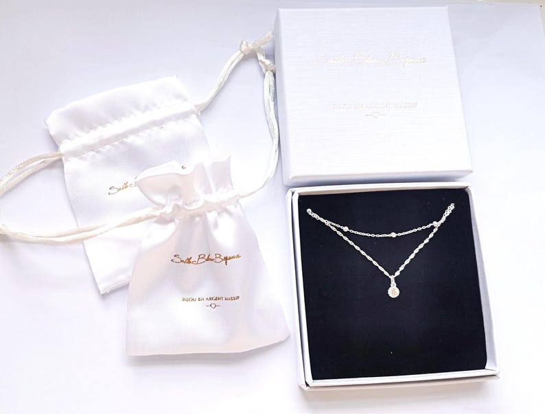 May include: A silver necklace with a small round pendant, packaged in a white box with a black interior and a white satin bag. The box and bag have the text "Solstice Blue Boutique" printed on them.