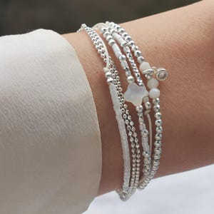 Op de afbeelding: Een set van vijf witte kralenarmbanden met zilveren accenten. De armbanden hebben een kleine witte sterbedel en een heldere kristallen bedel.