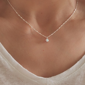 Könnte beinhalten: Eine zarte Silberkette mit einem kleinen, runden, weißen Opalanhänger. Die Halskette wird vor dem Ausschnitt einer Person gezeigt, mit einem weißen V-Ausschnitt-Shirt im Hintergrund. Die Kette ist fein und detailliert.