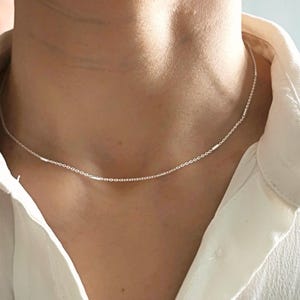 May include: A delicate silver chain necklace with small, rectangular links. The necklace is worn by a person, but the focus is on the necklace itself.
