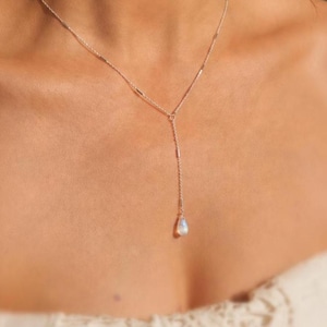 Op de afbeelding: Een zilveren lariat-ketting met een fijne ketting met kleine cilindrische metalen accenten en een enkele druppelvormige maansteen hanger die in het midden hangt. De ketting rust op het sleutelbeen met een Y-vormig ontwerp.