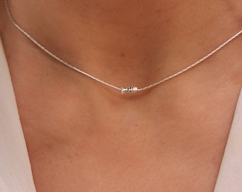 May include: A delicate silver necklace featuring a small fish pendant. The necklace is a thin, shimmering chain, and the fish pendant is centered. The necklace is worn around the neck, and the background is a white shirt.