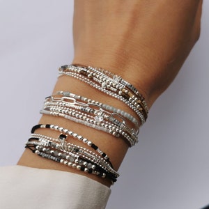 May include: A stack of silver and black beaded bracelets on a person's wrist. The bracelets are layered and feature different styles of beads, including small round beads, larger beads with a metallic finish, and beads with a chain-like design.