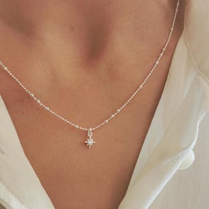 Könnte beinhalten: Eine zarte Silberkette mit einem kleinen Sternanhänger mit einem zentralen Edelstein. Die Kette hat eine feine Kette mit kleinen, gleichmäßig verteilten Perlen. Die Kette wird über einem weißen Hemd getragen.