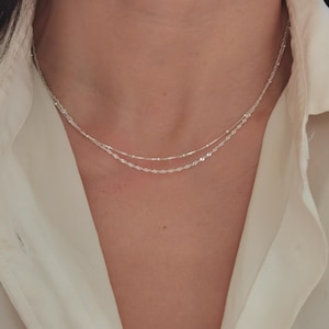 Pode incluir: Um close-up de um colar de prata com duas correntes finas. A corrente superior é uma corrente fina com pequenas contas, e a corrente inferior tem um design de corda torcida. O colar é usado contra uma camisa branca.