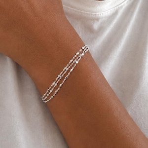 Op de afbeelding: Een drievoudige zilveren armband bestaande uit fijne kettinkjes met kleine, platte, reflecterende metalen schijfjes die gelijkmatig over elke streng zijn verdeeld. De armband heeft een heldere, gepolijste afwerking en een gelaagd ontwerp.