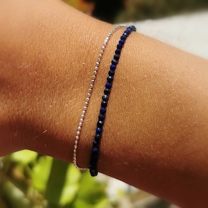 May include: Two delicate bracelets are shown on a wrist. One is a silver chain of small beads, and the other is a dark blue beaded bracelet. The bracelets are simple and elegant, suitable for everyday wear.