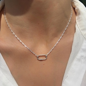 Peut inclure: Un collier argenté avec un pendentif rectangulaire. La chaîne est composée de petits maillons rectangulaires. Le pendentif est un rectangle plus grand et allongé. Le collier est porté sur une chemise blanche.