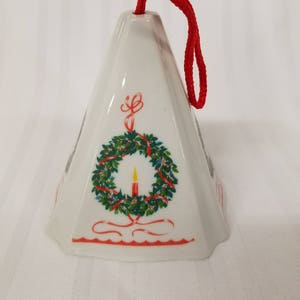 May include: White ceramic Christmas ornament shaped like a pyramid with a red ribbon hanger. The ornament features a green wreath with red ribbon and a yellow candle in the center.