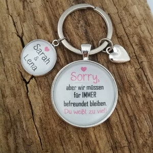 Könnte beinhalten: Ein silberner Schlüsselanhänger mit zwei runden Glasanhängern. Der erste Anhänger hat den Text "Sarah & Lena" mit einem rosa Herz. Der zweite Anhänger hat den Text "Sorry, aber wir müssen für IMMER befreundet bleiben. Du weißt zu viel!" mit einem rosa Herz.
