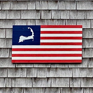 May include: A rectangular wooden sign featuring a red and white striped pattern with a blue canton on the left side. The blue section contains a white silhouette of the Cape Cod peninsula. The sign is mounted on a gray shingled exterior wall.